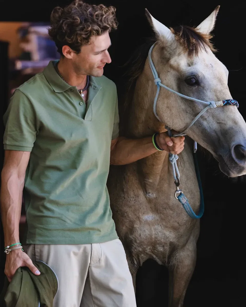 Hombre con polo verde junto a un caballo, primer plano de la muñeca con pulsera de cuentas y cordón.