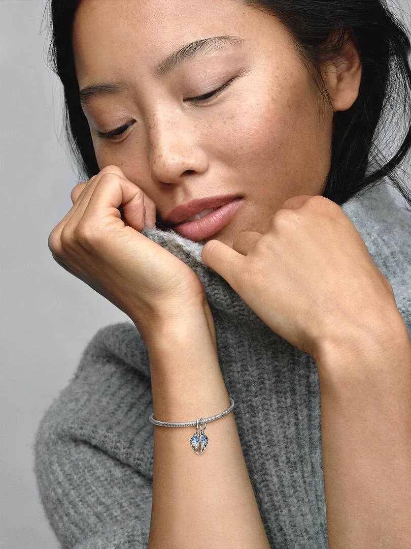 Gros plan du charm ailes bleu sur un bracelet au poignet, modèle en pull gris, fond neutre.