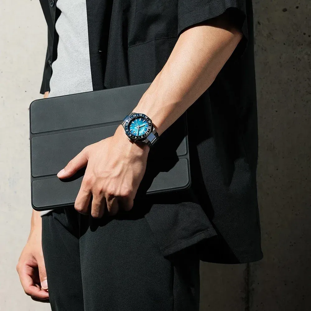 Modelo masculino con el reloj puesto en la muñeca, sosteniendo una tablet negra frente a una pared de hormigón
