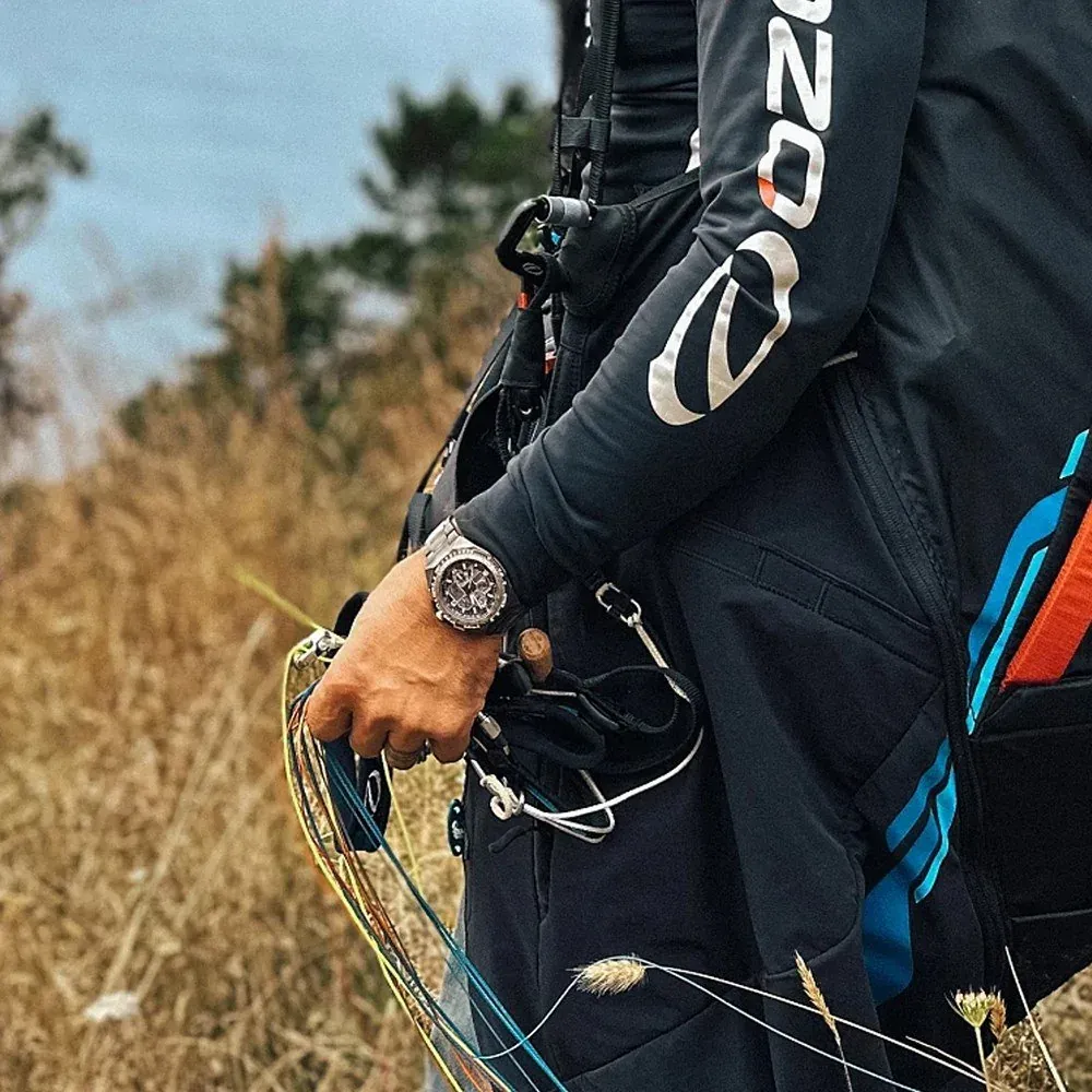 Reloj puesto en muñeca con ropa técnica negra y equipo de escalada, fondo de naturaleza desenfocado.