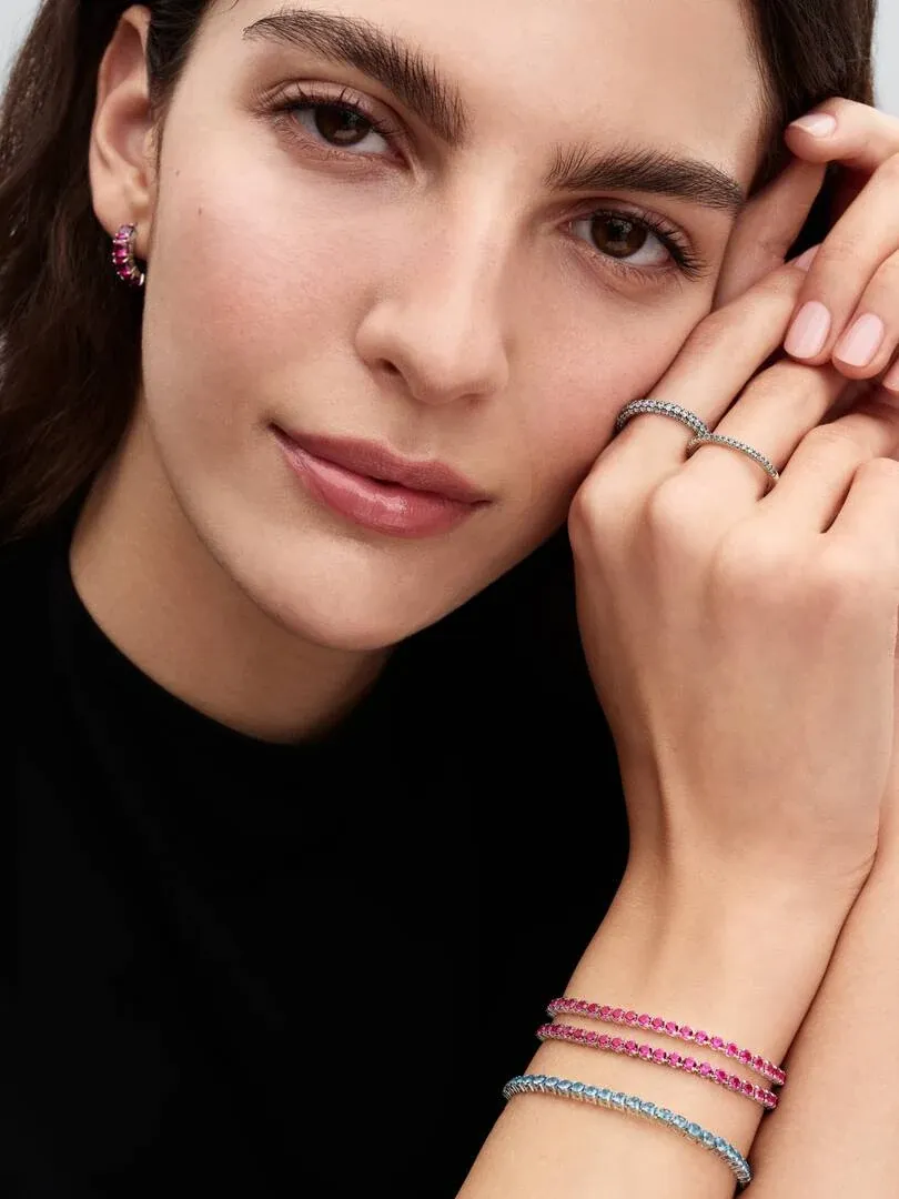 Primer plano de mano y muñeca con pulseras tenis rosa y azul, anillos finos y pendientes, sobre fondo oscuro.