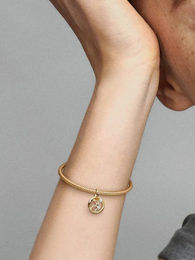 Close-up van de Virgo charm aan een gouden armband, gedragen om de pols tegen een neutrale grijze achtergrond.