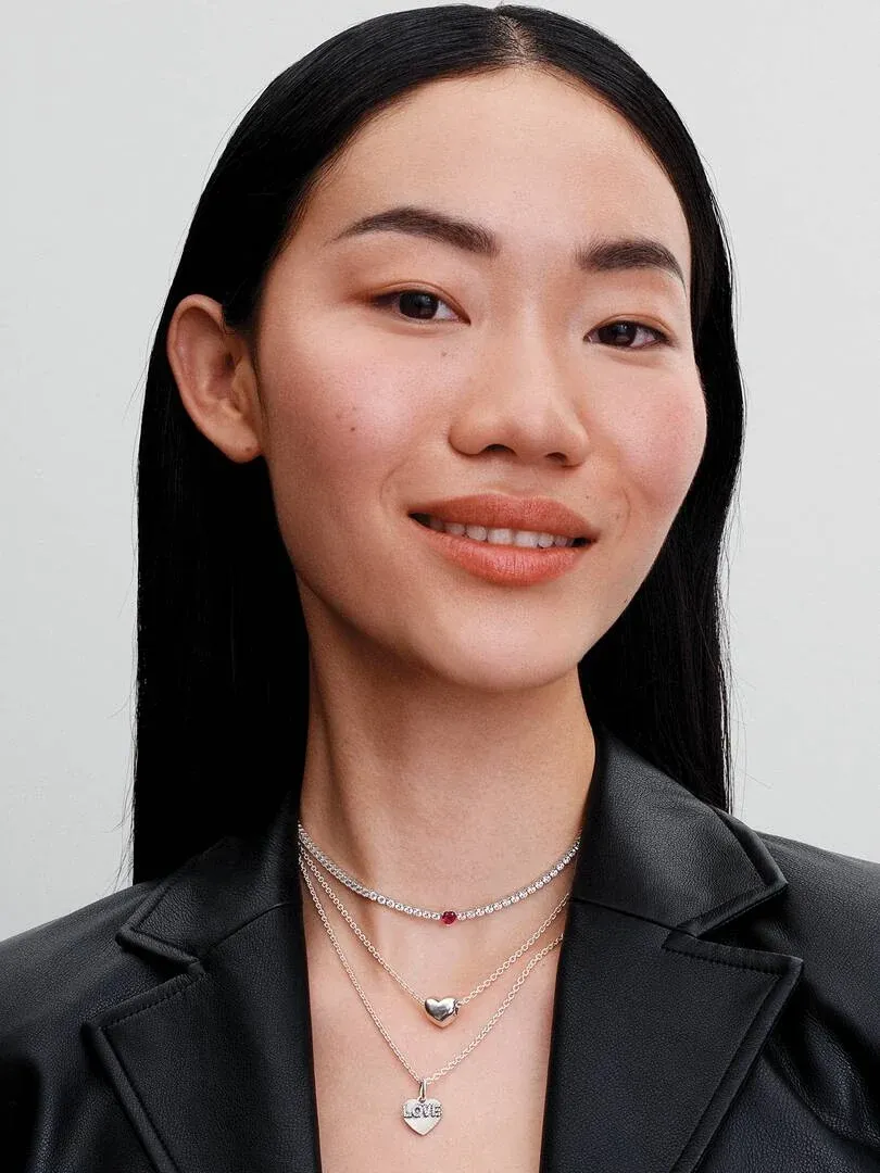 Primer plano de modelo luciendo tres collares de plata en capas, sobre chaqueta negra, fondo neutro.