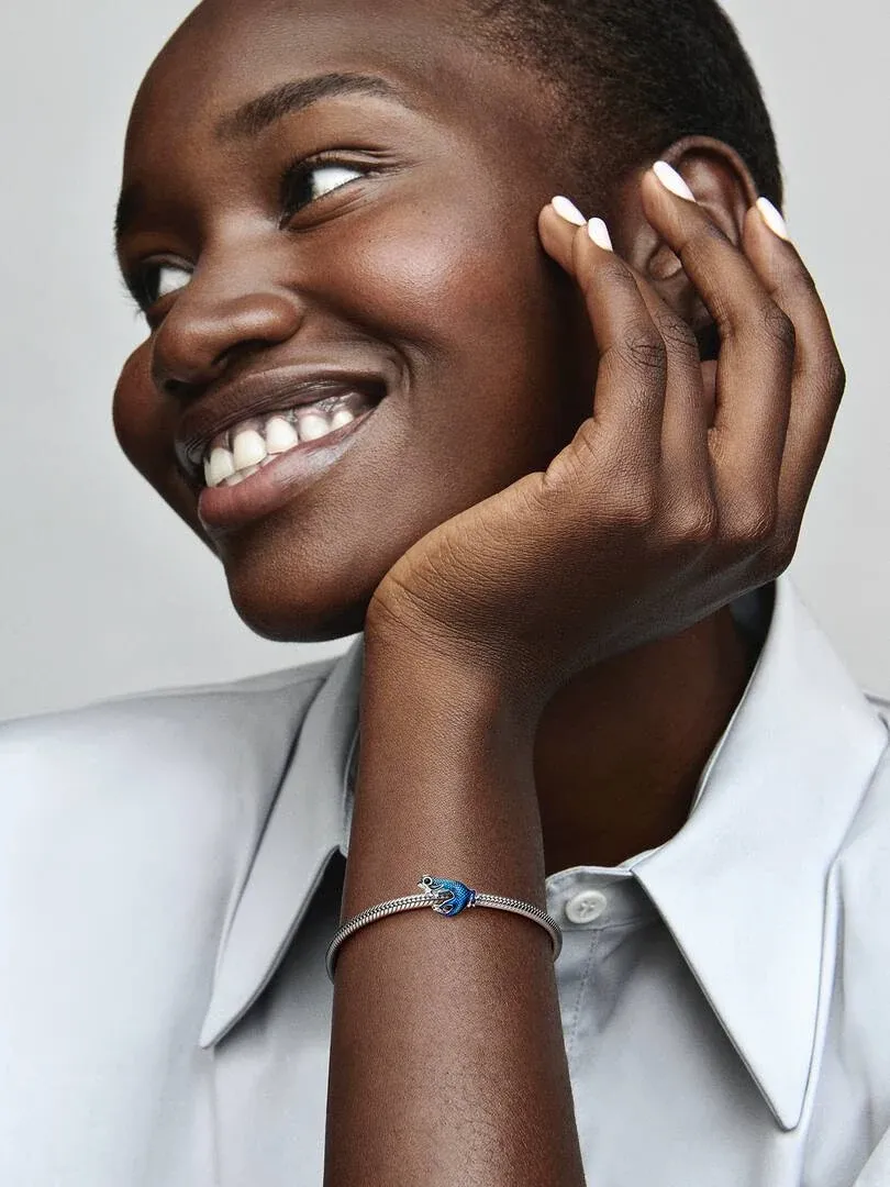 Primer plano de la muñeca con el charm de geco azul, modelo sonriente con camisa blanca, fondo neutro.