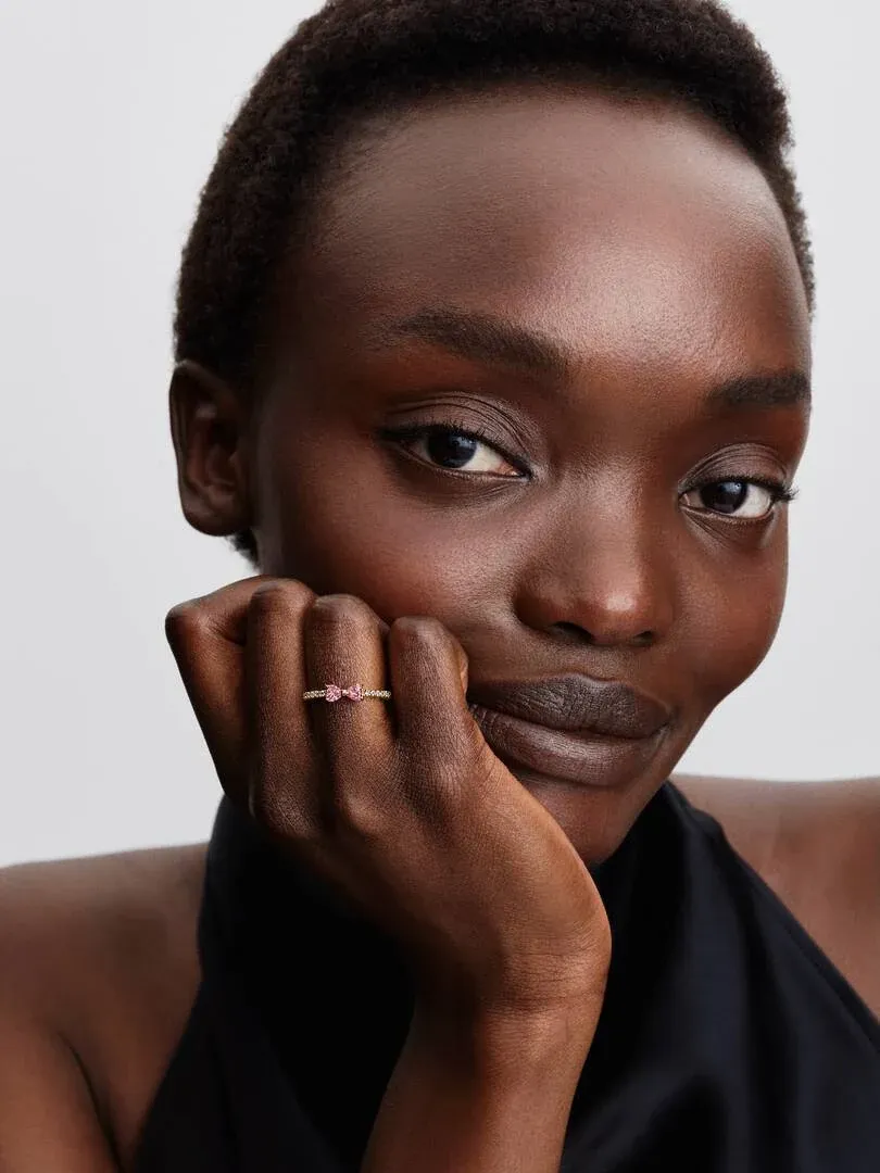 Primer plano de mano femenina apoyada en la cara, mostrando el anillo con gemas rosas sobre fondo blanco.
