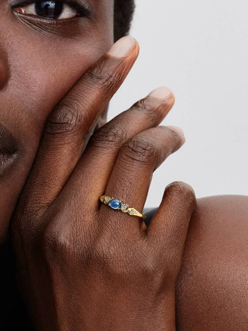 Primer plano lateral de un anillo dorado con gema azul en el dedo anular, sobre piel con fondo neutro.