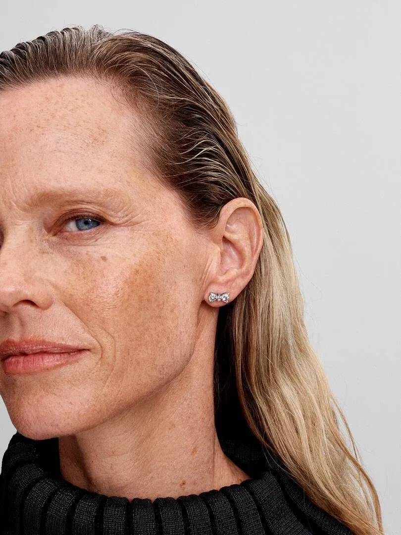 Gros plan d'une boucle d'oreille nœud étincelante portée sur le lobe de l'oreille, vue de profil sur fond neutre.