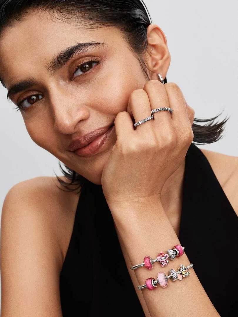 Primer plano de una mujer con pulsera de charms y anillo, toma lateral apoyada en la cara sobre fondo blanco.