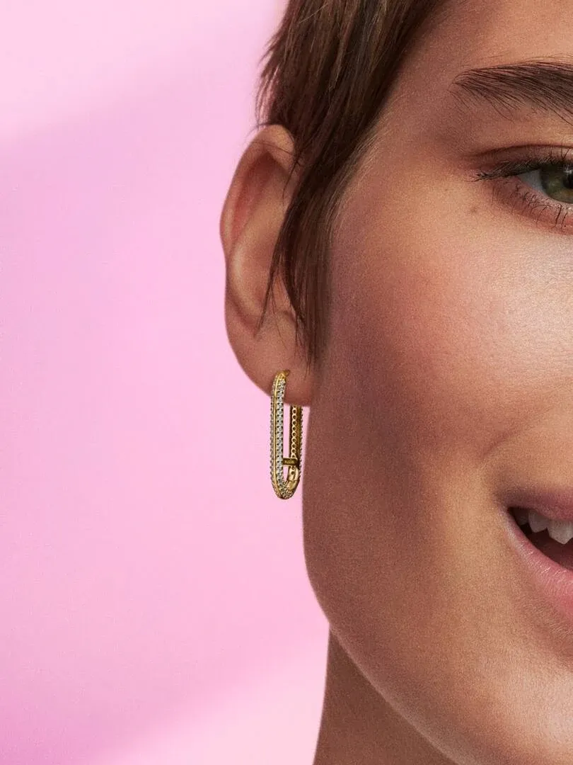 Side close-up of a rectangular pavé hoop earring worn on an ear against a pink background.