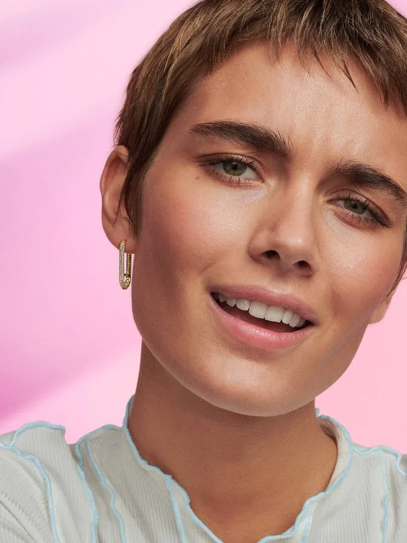 Close-up of a gold-plated rectangular pavé hoop earring worn on the ear, soft pink background.