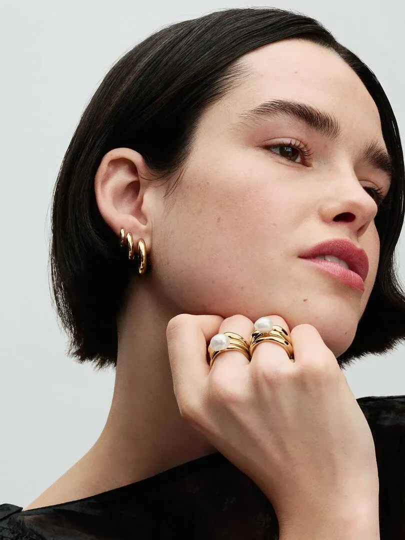 Primer plano de modelo con anillos de perla y pendientes de aro dorados, toma lateral sobre fondo neutro.