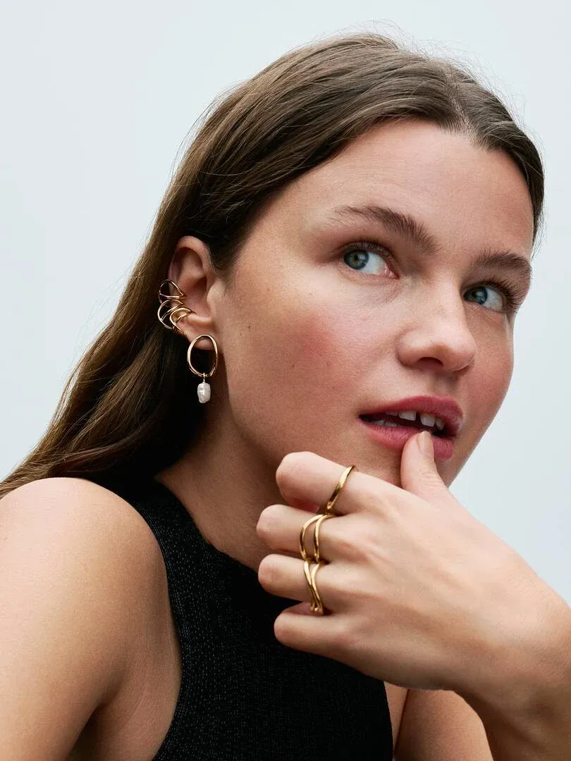 Primer plano de modelo con pendientes de aro dorados y perla, y anillos de banda orgánica en los dedos.
