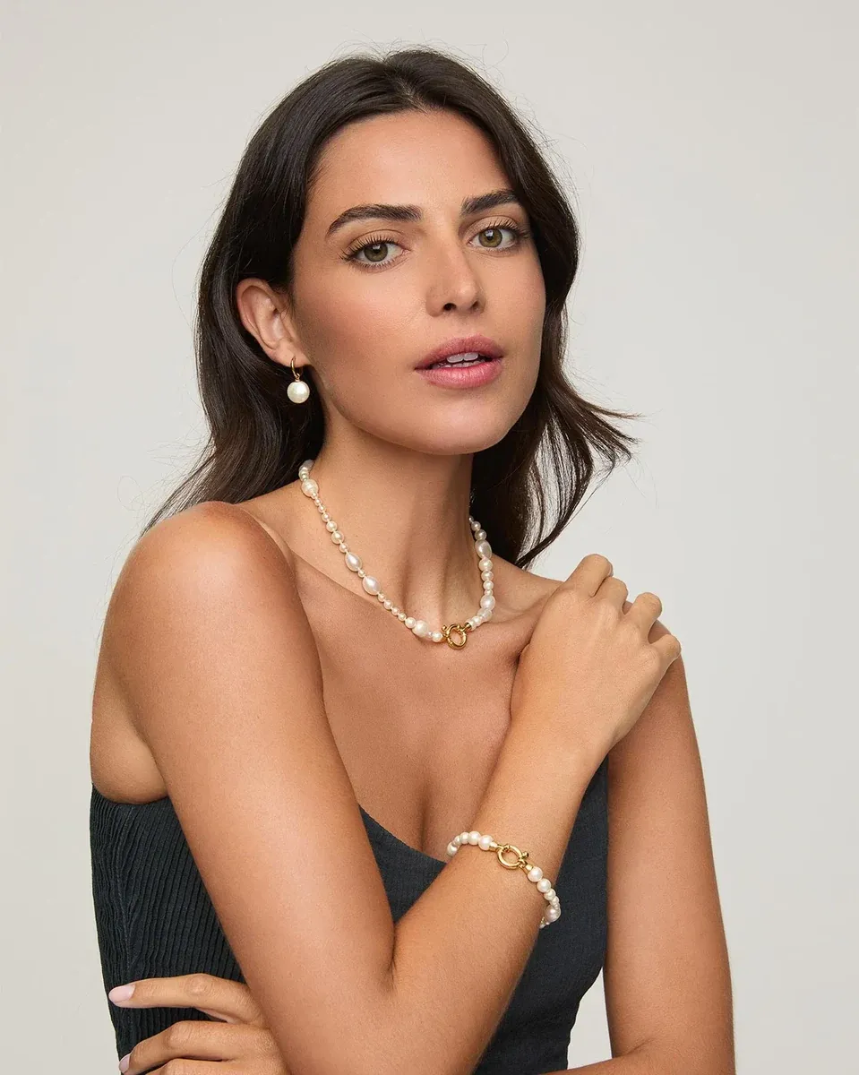 Model wearing pearl necklace and bracelet, close-up of face and torso against a neutral background.