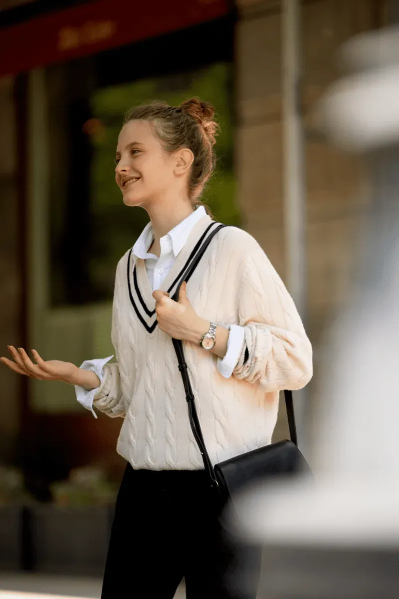 Plano medio de mujer con reloj en la muñeca, vestida con jersey blanco y bolso negro, fondo urbano desenfocado.