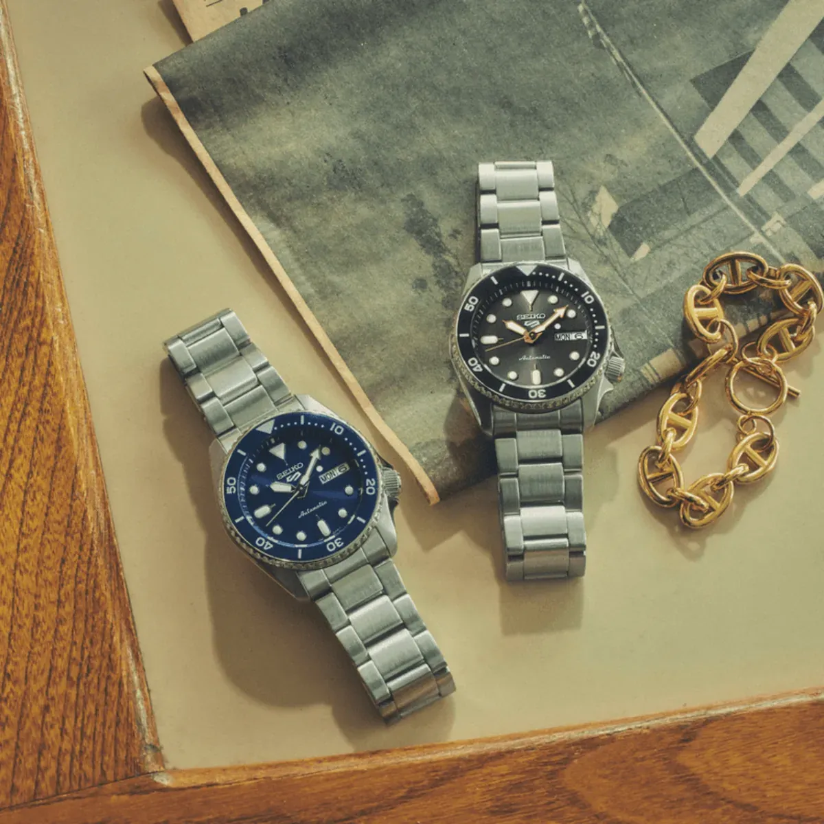 Composición cenital de dos relojes con esferas azul y negra y una cadena dorada sobre fondo neutro y revista.