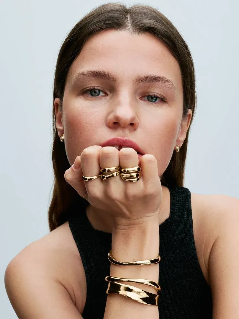 Primer plano de mano con pulsera rígida y anillos dorados, modelo posando frente a fondo neutro.