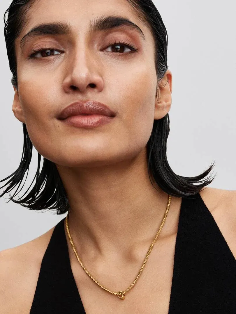 Close-up van een ketting met een letter J hanger op de hals van een model, neutrale grijze achtergrond.