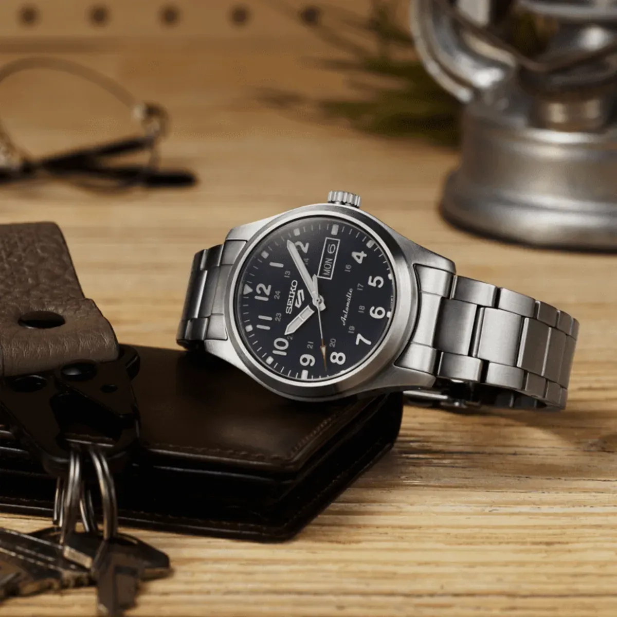Reloj apoyado sobre una mesa de madera junto a una cartera de cuero y llaves, fondo desenfocado.