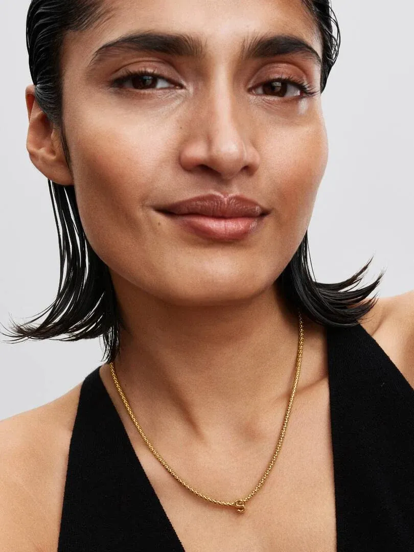 Nahaufnahme einer Halskette mit Buchstaben-Anhänger S am Hals eines Models, heller neutraler Hintergrund.