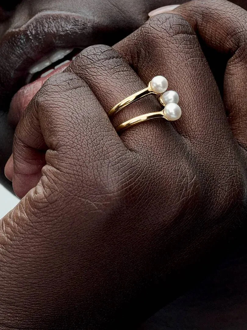 Primer plano lateral de anillo de tres bandas con perlas blancas en un dedo, sobre piel oscura.