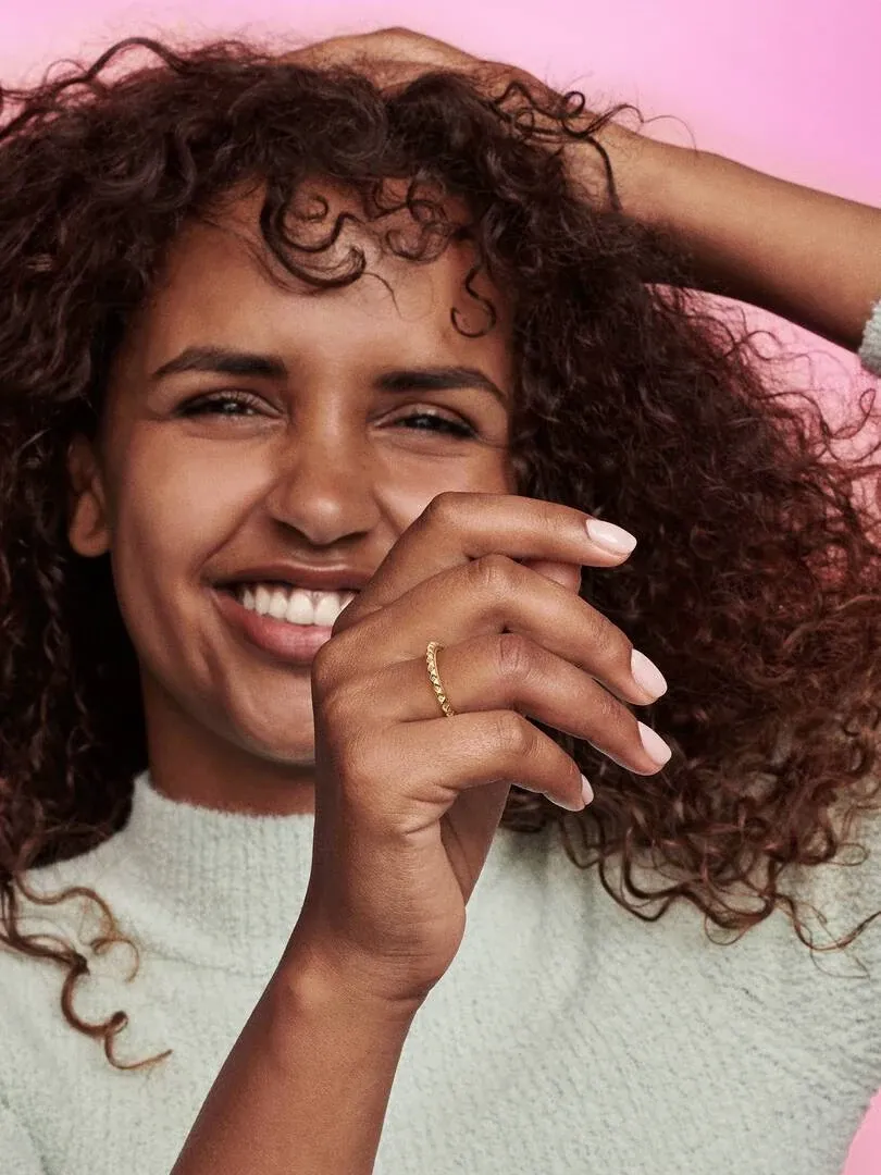 Primer plano de anillo dorado en dedo anular, mano femenina sobre fondo rosa, modelo sonriendo.