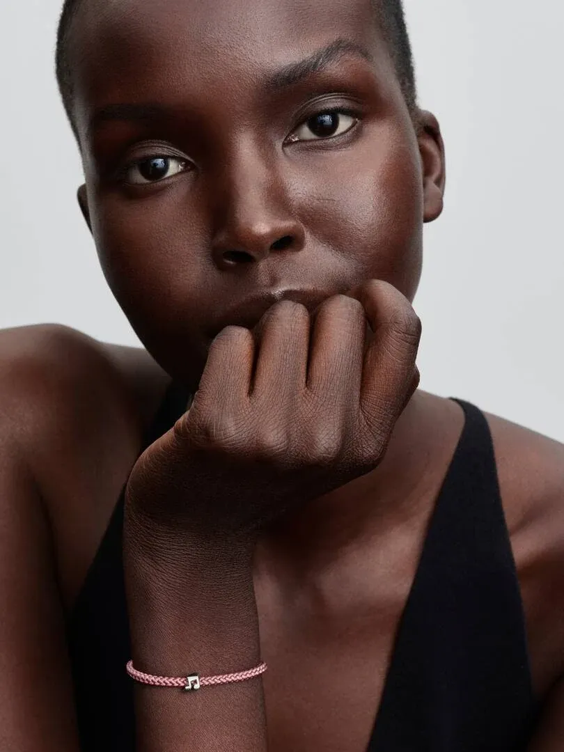 Primer plano de la muñeca con pulsera trenzada rosa y charm de nota musical, modelo con fondo neutro.