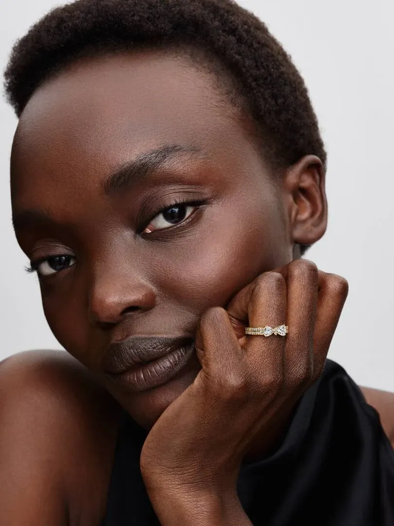 Primer plano del anillo de lazo dorado en el dedo, modelo apoyando la cara sobre su mano, fondo neutro.