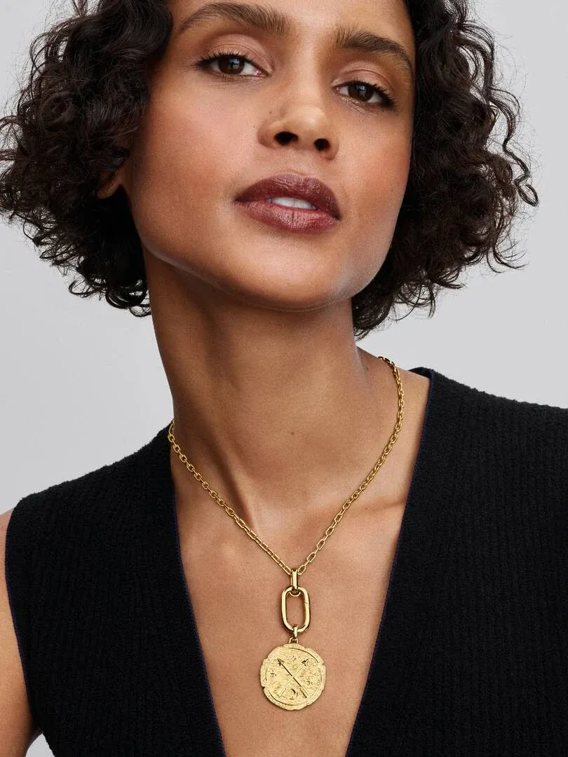 Close-up van de gouden medaillon op de hals van een model, vooraanzicht tegen neutrale achtergrond.