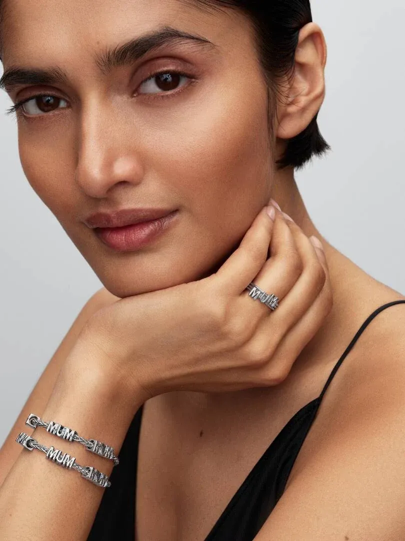 Nahaufnahme eines Models mit 'MUM' Charms am Handgelenk und einem passenden Ring, neutraler Hintergrund.
