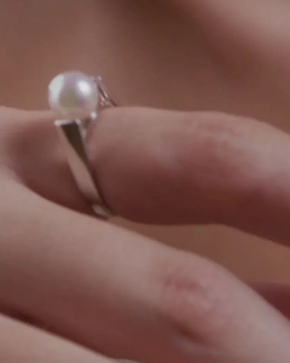 Side close-up of the white pearl ring worn on a finger, blurred background.