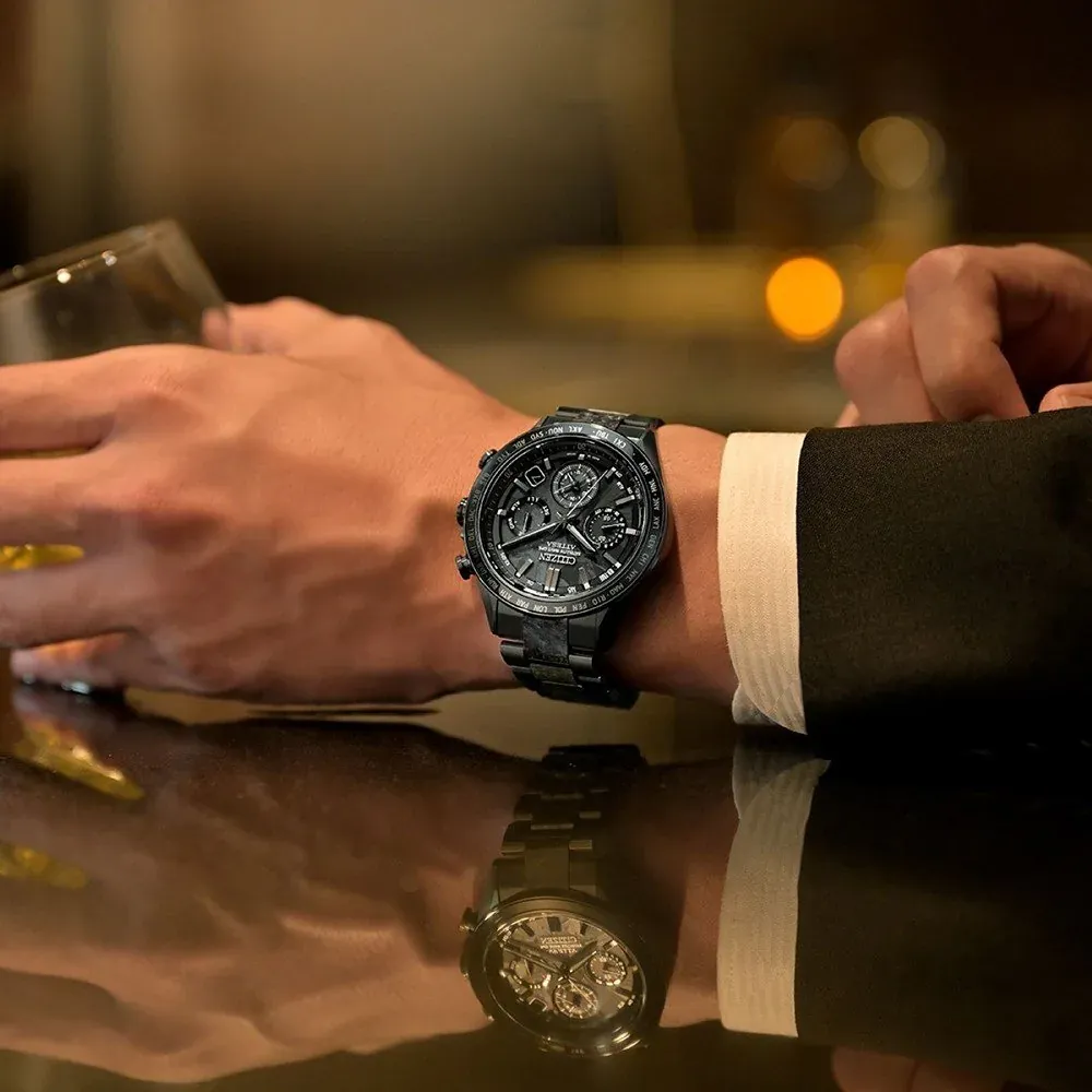 Reloj puesto en muñeca masculina, sosteniendo una copa sobre superficie reflectante, fondo desenfocado.