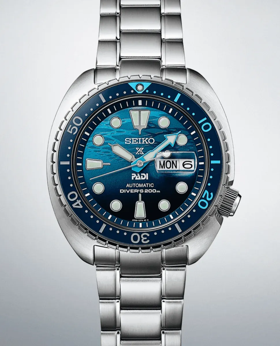 Vista frontal centrada del reloj con esfera azul y brazalete de acero, sobre fondo neutro degradado.