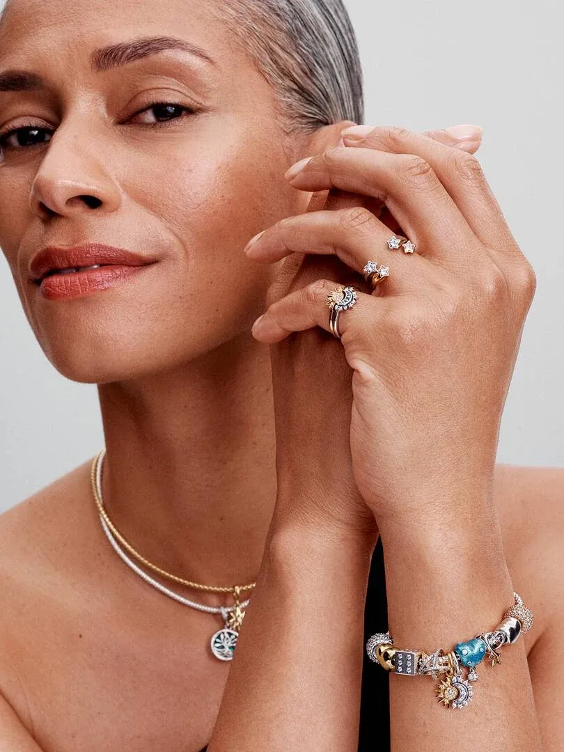 Gros plan d'un modèle portant un collier et un bracelet à charms, mains près du visage sur fond neutre.