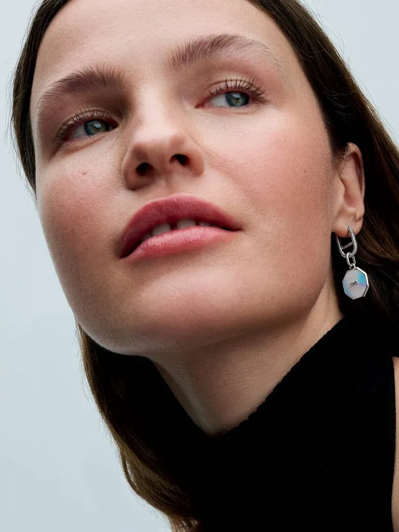 Gros plan latéral d'une boucle d'oreille créole avec pendentif hexagonal portée sur l'oreille, fond neutre.