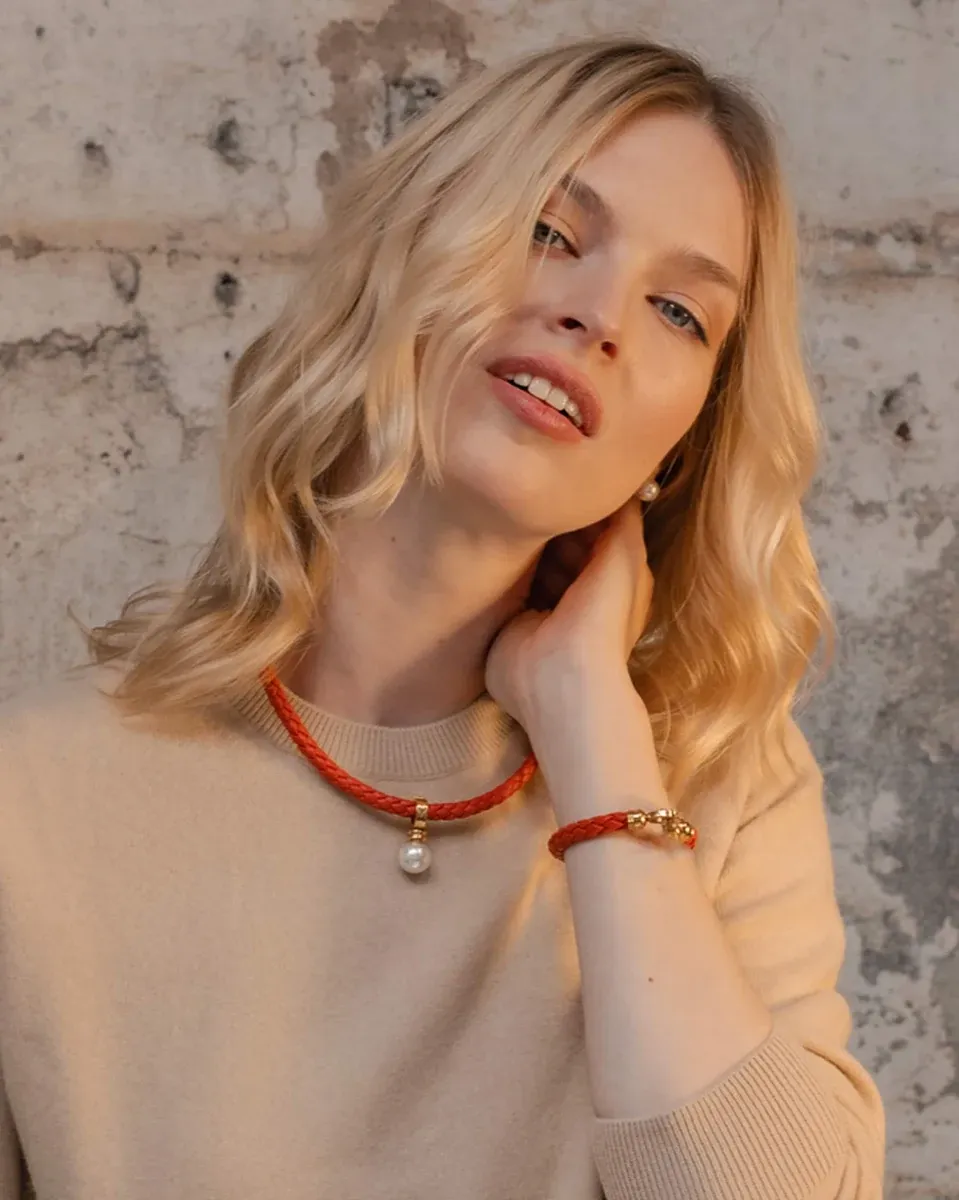 Modelo con collar de cuero naranja y perla, toma frontal media sobre fondo de textura rústica.