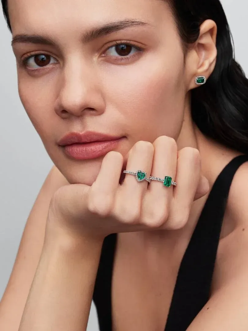 Gros plan d'un modèle portant des boucles d'oreilles vertes et une bague halo, vue de face sur fond neutre.