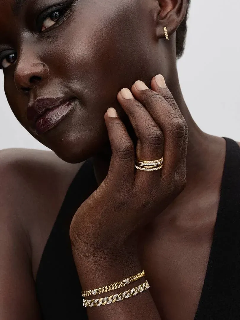 Primer plano de la mano con el anillo y pulseras doradas, modelo con piel oscura sobre fondo neutro.