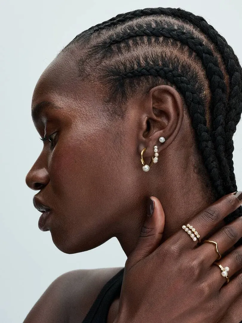 Primer plano de perfil con pendientes de perlas y anillo de oro en la mano, fondo neutro claro.