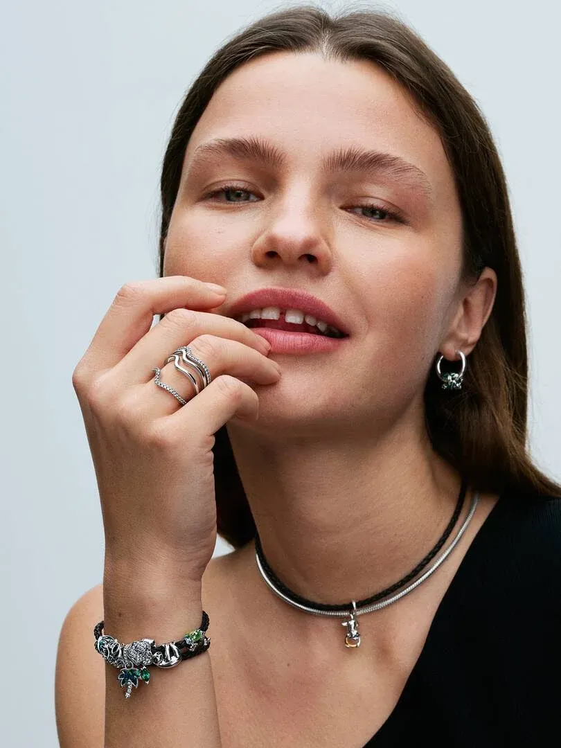 Primer plano de modelo luciendo pulsera de cuero y collar con charms, toma frontal con mano cerca del rostro.