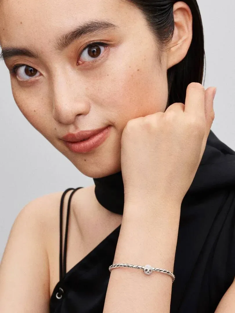 Primer plano de la muñeca con pulsera de plata y charm de calavera, modelo con top negro sobre fondo gris.