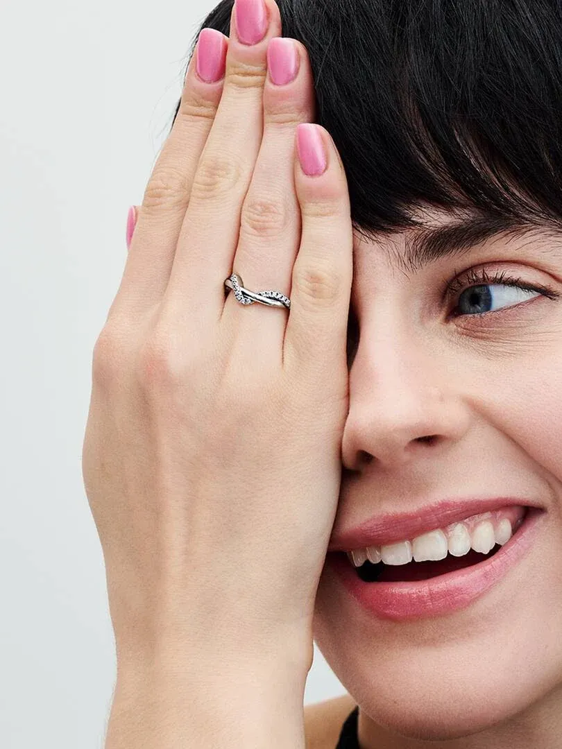 Nahaufnahme eines wellenförmigen Pavé-Rings am Ringfinger, Hand am Gesicht, neutraler Hintergrund.