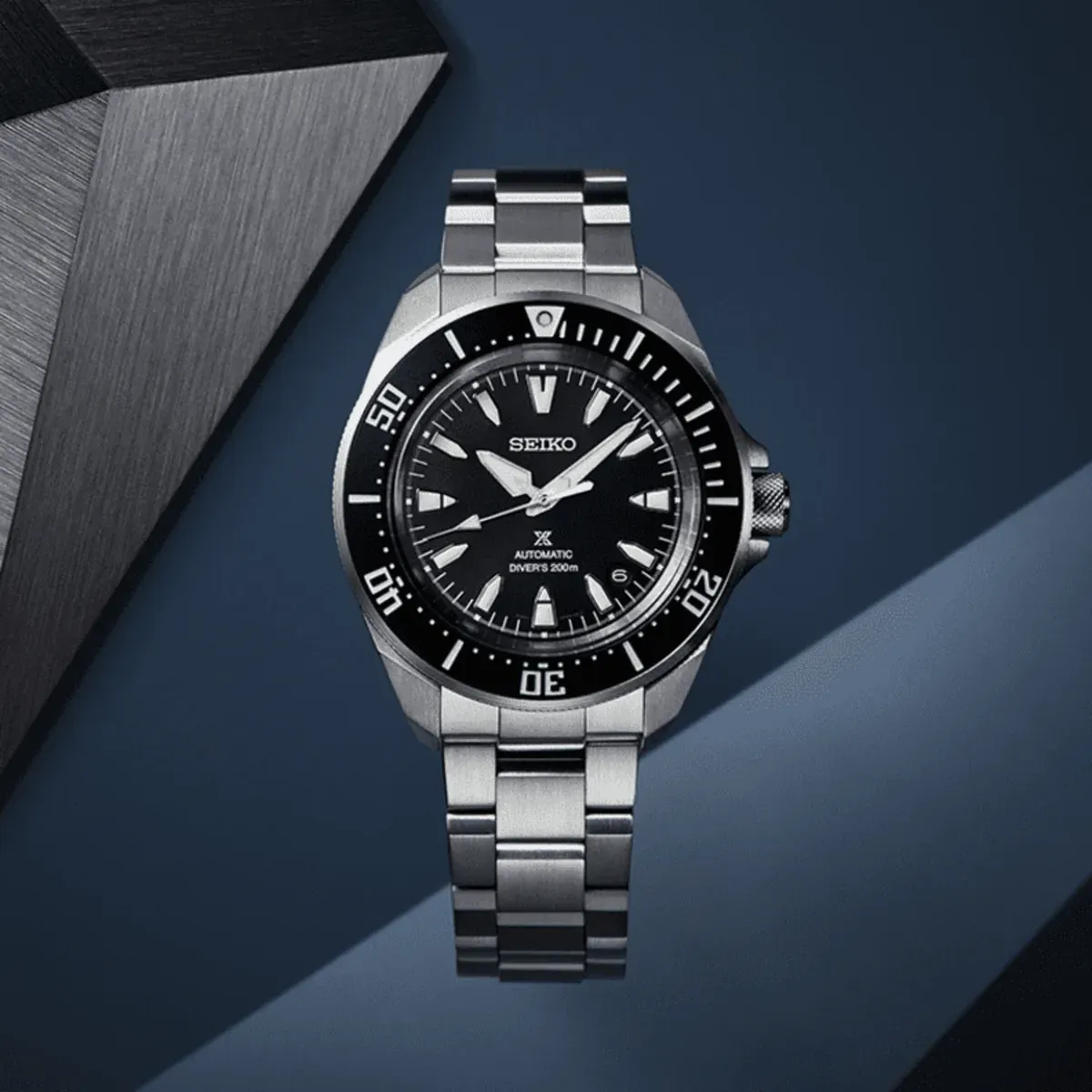 Reloj visto frontalmente sobre fondo azul oscuro con elemento geométrico metálico y luz contrastada.