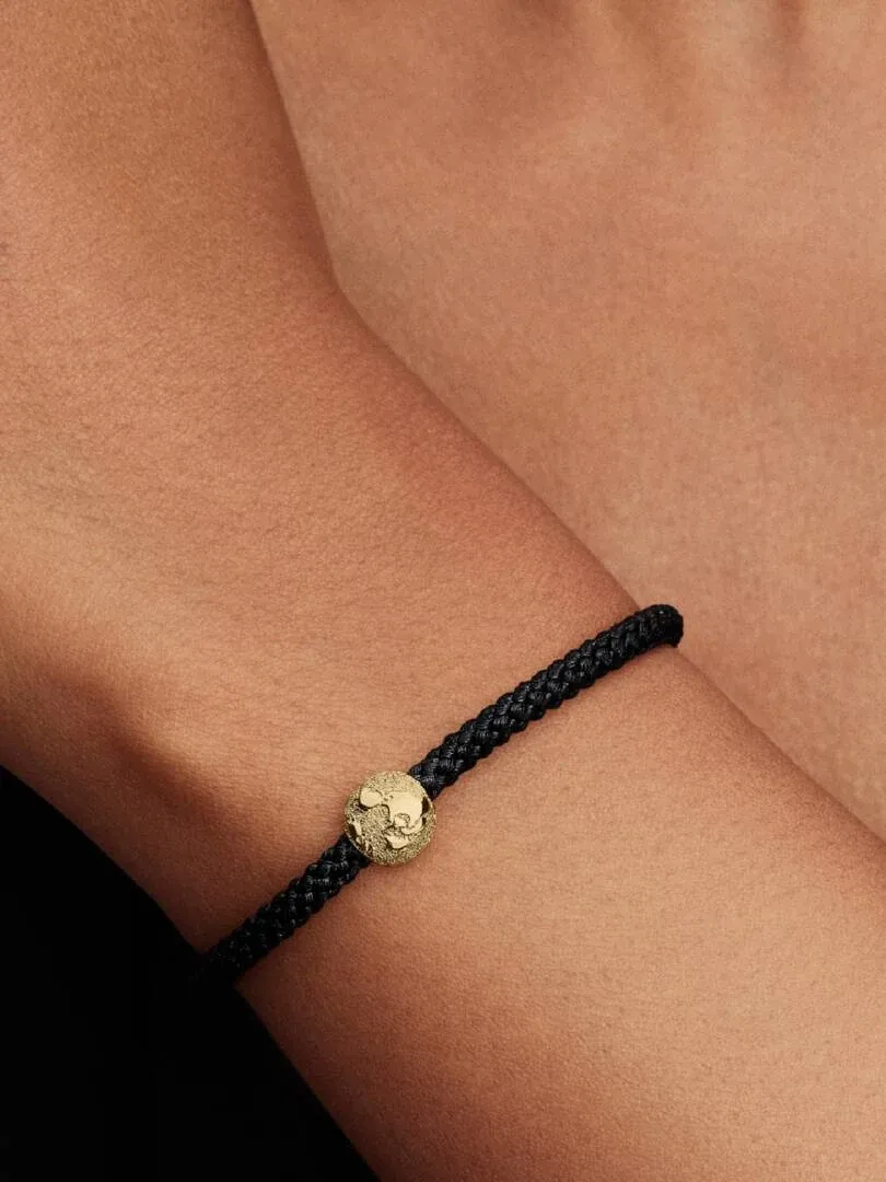 Primer plano del charm de globo terráqueo dorado en una pulsera negra, sobre la muñeca.