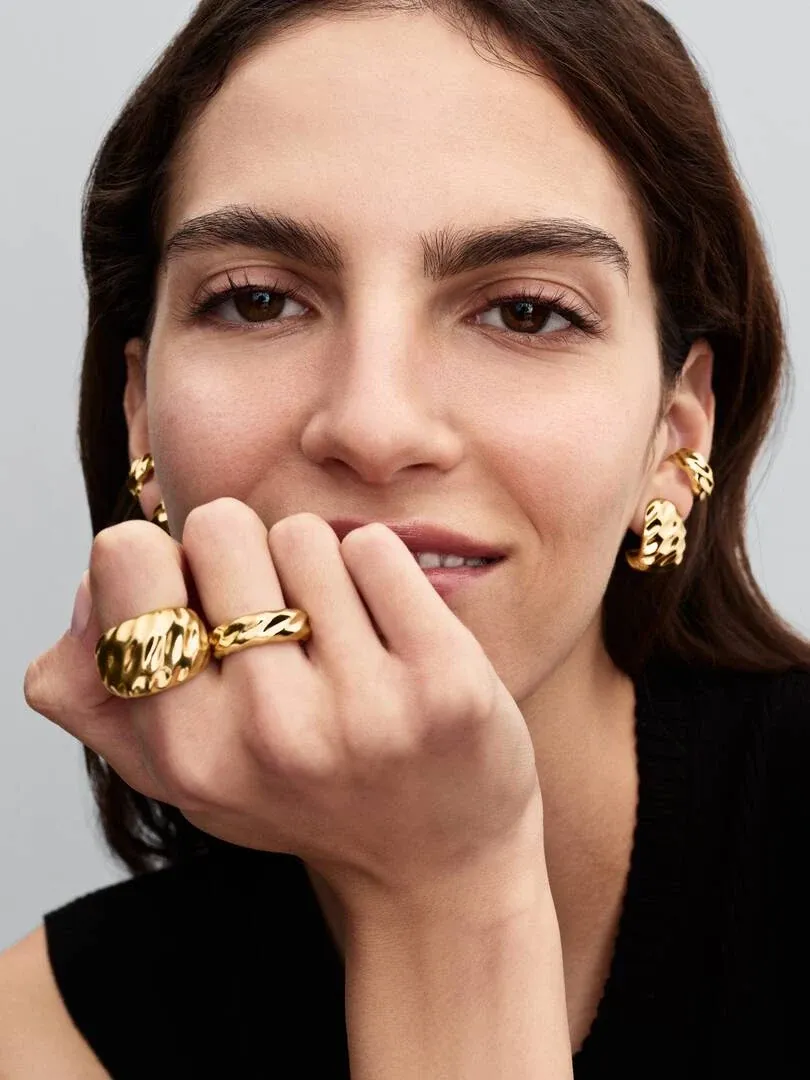 Primer plano de mano femenina con anillo dorado ondulado, modelo posando frente a fondo neutro.