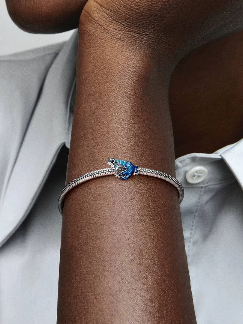 Primer plano del charm de geco azul sobre una pulsera de plata, puesto en la muñeca con camisa blanca.