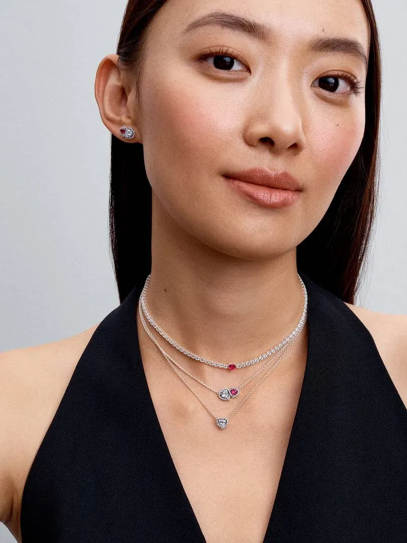 Gros plan d'une femme portant des colliers superposés avec cœurs rouges et brillants, fond neutre.