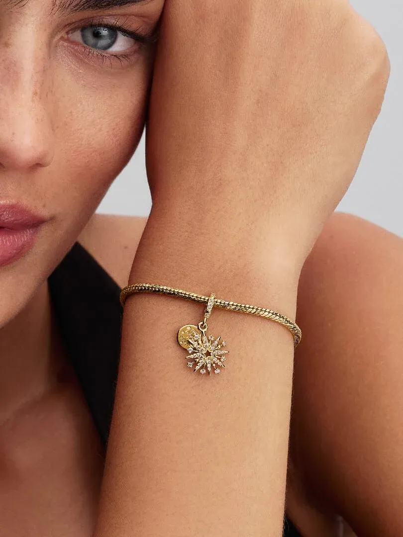 Close-up van dubbele charm aan een gouden armband, gedragen om de pols tegen een wazige achtergrond.