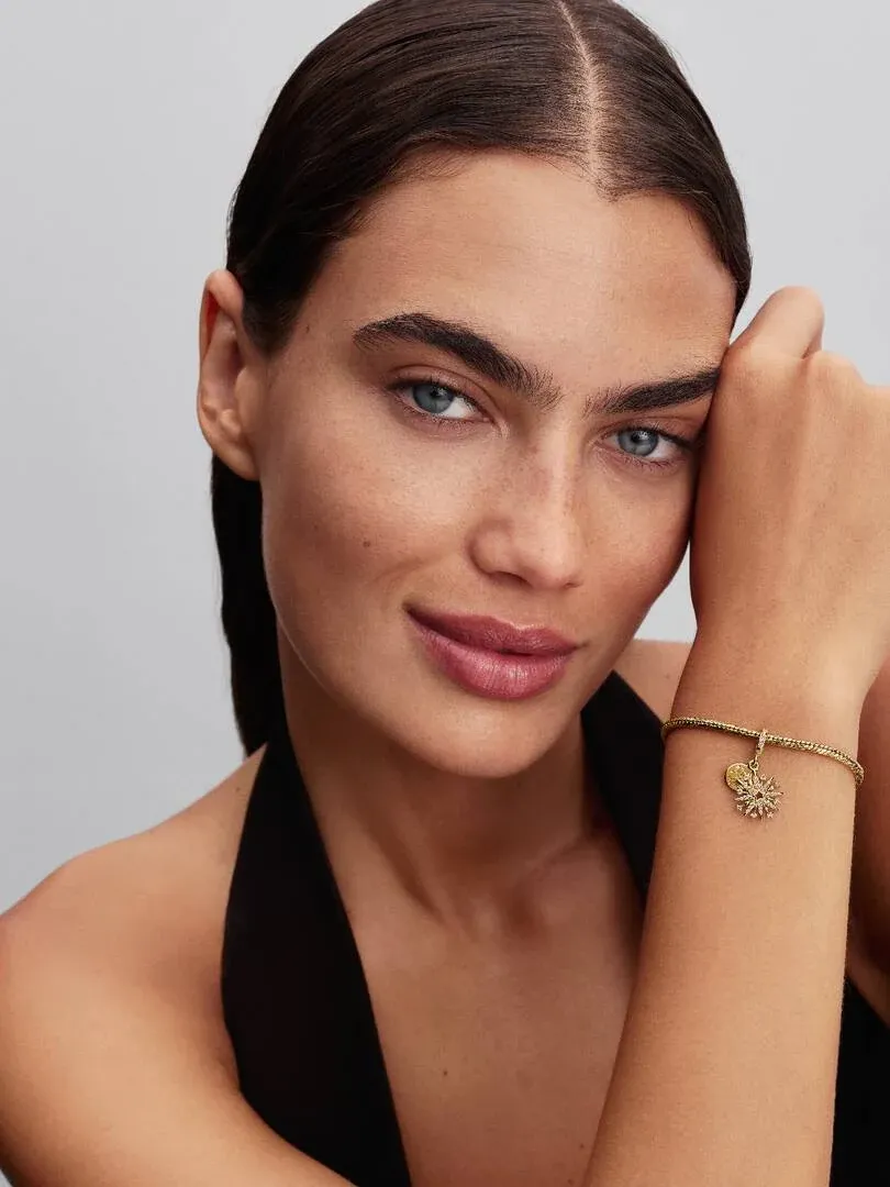 Close-up van de gouden charm om de pols, zijaanzicht op een model met neutrale achtergrond.