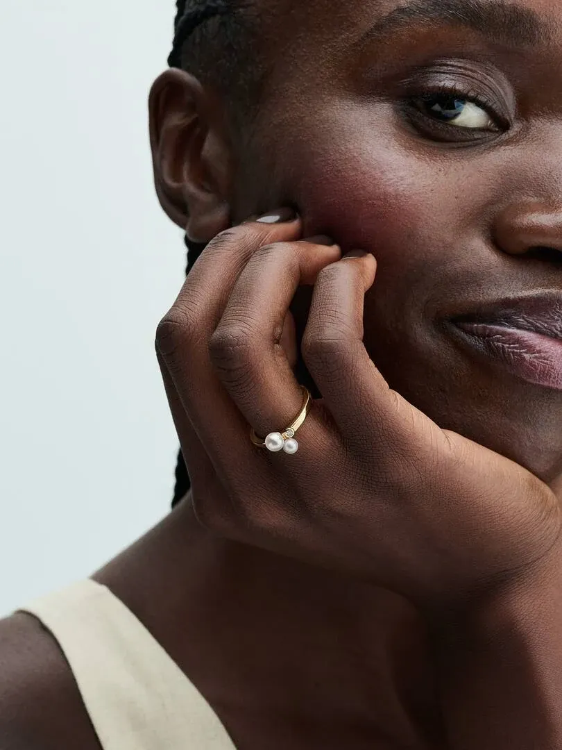 Primer plano del anillo con perlas en el dedo anular, modelo apoyando la mano en la cara sobre fondo neutro.