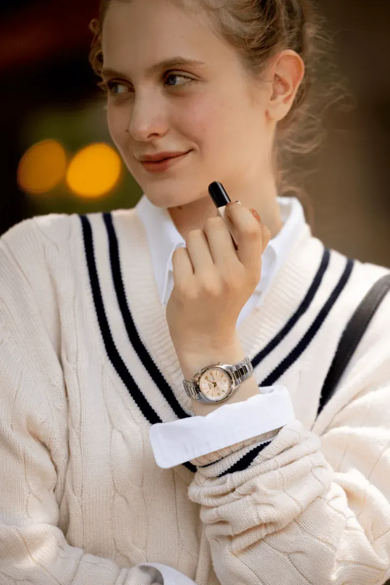Mujer con reloj en la muñeca, vistiendo jersey blanco de punto, fondo exterior desenfocado.